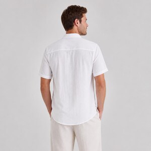 May include: A short-sleeved white button-down shirt with a textured fabric finish, featuring a classic collar and a straight back yoke. The shirt is paired with light beige trousers, showcasing a clean and simple design from the back view.