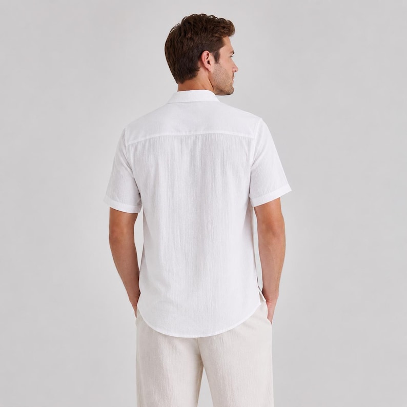 May include: A short-sleeved white button-down shirt with a textured fabric finish, featuring a classic collar and a straight back yoke. The shirt is paired with light beige trousers, showcasing a clean and simple design from the back view.