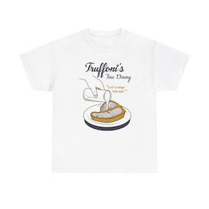 Può includere: T-shirt bianco con un'illustrazione a cartoni animati di una bistecca a cui viene versato un liquido bianco. Il testo "Truffoni's Fine Dining" è sopra l'immagine, con la frase "Let's slop 'em up!" sotto.