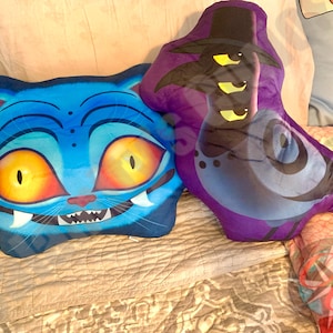 May include: Two plush pillows. One is blue and cat-shaped with orange eyes and a wide grin. The other is purple with a hat and three yellow eyes. Both are on a bed with a patterned comforter.
