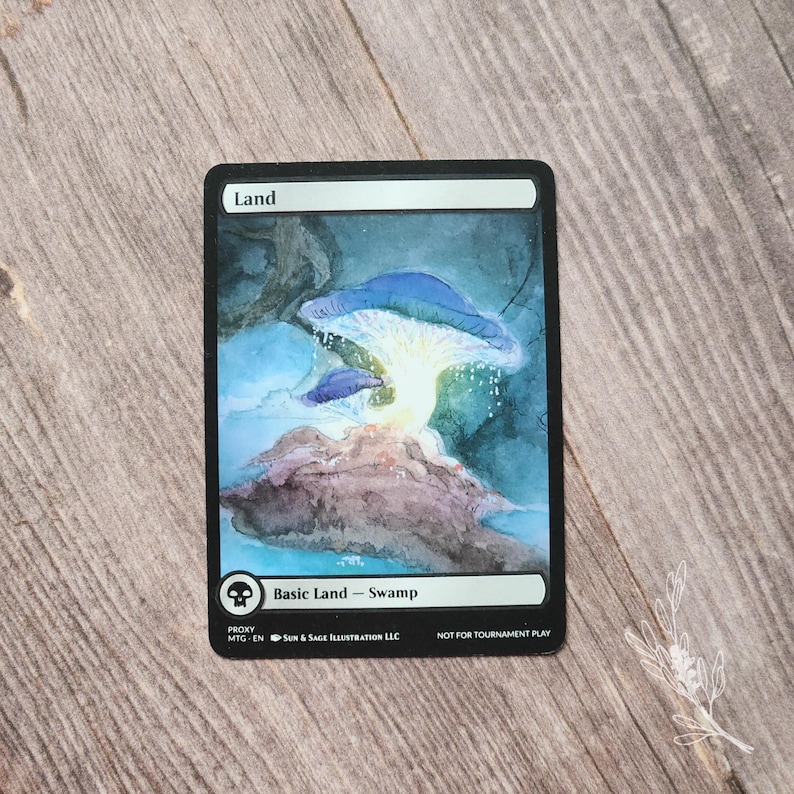 May include: A proxy Magic: The Gathering card featuring a watercolor illustration of a swamp scene. The card has a black border and displays a blue mushroom with a glowing center. Text includes "Land" and "Basic Land - Swamp."