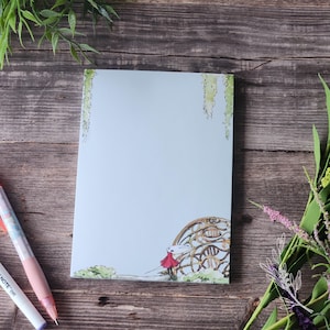 May include: A light blue notepad with an illustration of a figure in a red cloak and a decorative architectural element. The notepad is surrounded by pens and greenery on a wooden surface.