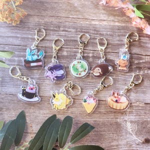 May include: Collection of clear acrylic keychains with gold-colored metal hardware. The keychains feature various cartoon character designs, including a blue creature on a cake, a purple creature with a macaron, and a yellow creature.