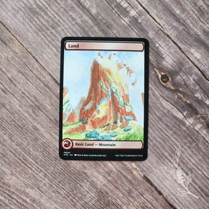 May include: A Magic: The Gathering proxy card featuring a watercolor illustration of a mountain landscape. The card has a black border and text that reads "Land" at the top and "Basic Land - Mountain" at the bottom. The artwork depicts a brown mountain with blue and green accents.