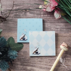 May include: Two light blue square sticky note pads. One pad has a diamond pattern, the other has a diamond pattern and a cartoon ghost with a quill and inkwell. A pen with a cat-shaped top is in the image.