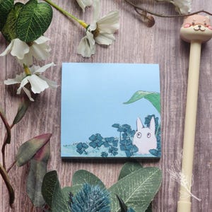 May include: A square, light blue notepad featuring a cartoon rabbit peeking from behind blue clovers and a green leaf. A pen with a dog-like character on top is visible. Decorative white flowers and green leaves surround the notepad.