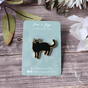 May include: A black cat enamel pin with gold accents. The pin is on a blue card with the text "Sun + Sage Illustration" and social media handles.