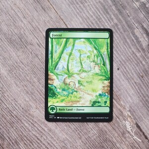 May include: A rectangular card with a green border and the word "Forest" at the top. The card features a watercolor painting of a forest scene with green foliage and a path. The text "Basic Land - Forest" is at the bottom.