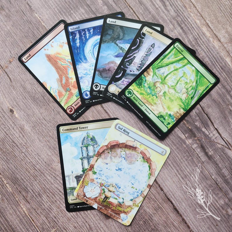 May include: A collection of Magic: The Gathering trading cards featuring various landscape illustrations. The cards include "Island," "Land," "Forest," "Command Tower," and "Sol Ring." The artwork showcases watercolor-style paintings of natural environments.