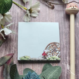 May include: A light blue square sticky note with an illustration of a character in a red cape and a decorative brown archway. A beige pen with a dog-shaped topper is on the right. White flowers and green leaves are in the background.