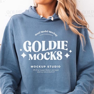 Puede incluir: Una sudadera con capucha azul claro con el texto "GOLDIE MOCKS" en blanco. La sudadera tiene un corte holgado y la lleva puesta una persona sentada en un sofá blanco.