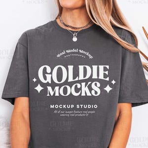 Może przedstawiać: Szara koszulka z nadrukiem "GOLDIE MOCKS" i "MOCKUP STUDIO" w kolorze białym. Koszulka jest noszona przez osobę. Koszulka ma luźny krój i krótkie rękawy. Obraz zawiera również naszyjnik.