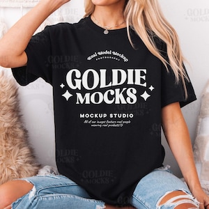 Op de afbeelding: Een zwart T-shirt met de tekst "GOLDIE MOCKS" in wit. Het shirt wordt gedragen door een persoon die op een wit en bruin pluche oppervlak zit. De tekst "Real Model Mockup" staat boven de tekst "GOLDIE MOCKS". De tekst "MOCKUP STUDIO" staat onder de tekst "GOLDIE MOCKS".