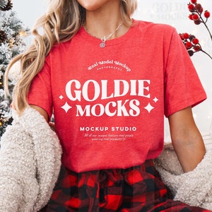 May include: A red t-shirt with white text that reads "Goldie Mocks" and "Mockup Studio". The shirt is being modeled by a person wearing red and black plaid pants.