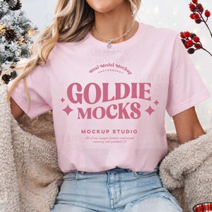 May include: A pink t-shirt with the text "GOLDIE MOCKS MOCKUP STUDIO" in a pink retro font. The shirt is being worn by a person with a white fuzzy cardigan and blue jeans.
