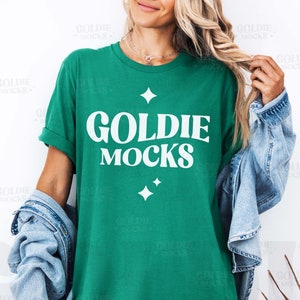 May include: A green t-shirt with the text "Goldie Mocks" in white lettering. The shirt has a relaxed fit and is being modelled by a person wearing a denim jacket.