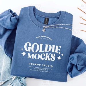 Può includere: Una felpa blu navy con la scritta bianca "GOLDIE MOCKS", abbinata a jeans strappati di colore chiaro e una collana d'argento. Una foglia di palma secca aggiunge un elemento naturale. La felpa è del marchio Gildan.