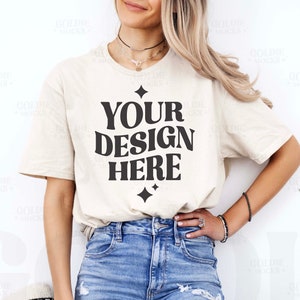 Puede incluir: Camiseta color crema con el texto "YOUR DESIGN HERE" en letras negras en negrita. La camiseta se combina con vaqueros de mezclilla azul claro. La modelo lleva joyas y tiene el pelo rubio.