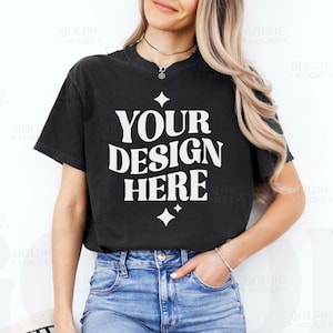 Puede incluir: Una camiseta negra con el texto "YOUR DESIGN HERE" en blanco. La camiseta la lleva puesta una persona con el pelo largo y rubio y está metida en unos vaqueros azules.