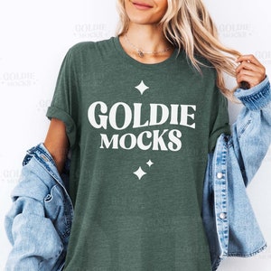 May include: A green t-shirt with the text "Goldie Mocks" in white lettering. The shirt is being worn by a person with long blonde hair and a denim jacket.