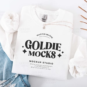 Puede incluir: Una maqueta de sudadera Gildan blanca con el texto "GOLDIE MOCKS" en negro. La sudadera está colocada sobre un par de vaqueros azules con detalles desgastados. También hay un collar plateado y hojas secas.