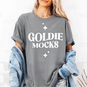 Peut inclure: Un t-shirt gris surdimensionné avec le texte "GOLDIE MOCKS" en lettres blanches. Le t-shirt a une coupe ample et présente un petit motif d'étoile.