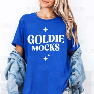 May include: A royal blue t-shirt with a white graphic that says "Goldie Mocks" with four white stars. The shirt is being worn by a person with long blonde hair and a denim jacket.