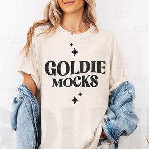 May include: A white t-shirt with a black graphic that says "Goldie Mocks" with stars on either side of the text.