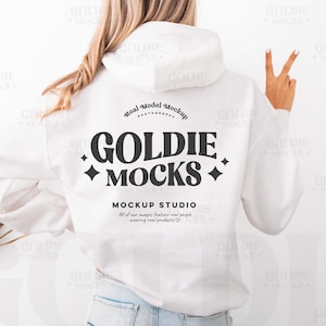 以下が含まれることがあります： 背中に黒い文字で「GOLDIE MOCKS」と書かれた白いパーカー。メインロゴの下のテキストが示すように、パーカーはモックアップスタジオのものです。モデルは水色のジーンズを履いています。