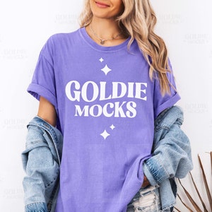 May include: A woman wearing a purple t-shirt with the text "Goldie Mocks" in white. The shirt has a distressed look and features small white stars.