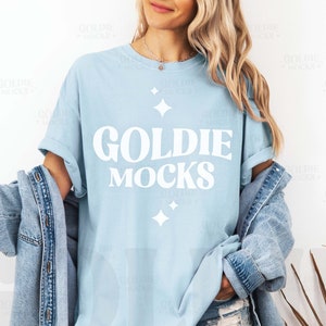 May include: A light blue t-shirt with the text "Goldie Mocks" in white lettering. The shirt has a relaxed fit and features a distressed look.