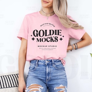 Puede incluir: Una camiseta rosa con el texto "GOLDIE MOCKS" en letras negras. El texto está rodeado de pequeños diamantes. El texto también incluye las palabras "real pod mockup" y "MOCKUP STUDIO". La camiseta la lleva puesta una persona con el pelo largo y rubio. La persona lleva unos vaqueros azules con detalles desgastados.