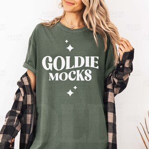 Puede incluir: Una camiseta verde con el texto "Goldie Mocks" en letras blancas. La camiseta tiene un corte holgado y cuello redondo.