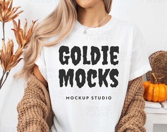 May include: A white t-shirt with a black graphic that says "Goldie Mocks Mockup Studio". The shirt is being worn by a person with long blonde hair. The person is wearing a brown cardigan and black pants.