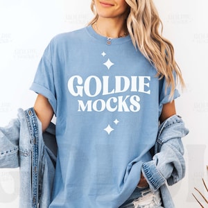 Può includere: Una maglietta blu chiaro con il testo "Goldie Mocks" in lettere bianche. La maglietta ha una vestibilità rilassata e un collo rotondo.