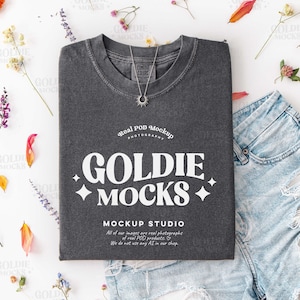 Puede incluir: Una camiseta gris oscuro con el texto "GOLDIE MOCKS" en blanco, junto con un collar plateado. La camiseta se combina con pantalones cortos de mezclilla y está rodeada de flores y pétalos coloridos, creando una composición plana.
