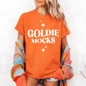 Puede incluir: Una mujer lleva una camiseta naranja con el texto "Goldie Mocks" en blanco. La camiseta tiene un corte holgado y cuello redondo. Lleva un cárdigan multicolor sobre la camiseta.