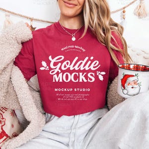 May include: A red t-shirt with the text "Goldie Mocks" and a Santa Claus mug. The mug has a red and black plaid interior and a Santa Claus graphic. The shirt also has additional text. The person is wearing a cream-colored cardigan.