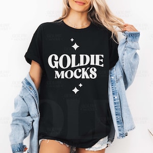 Puede incluir: Camiseta negra con el texto blanco "GOLDIE MOCKS" y gráficos de estrellas. Se lleva una chaqueta vaquera azul claro. La modelo se está tocando el pelo.