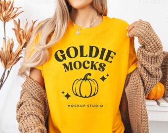 May include: A yellow t-shirt featuring the text "Goldie Mocks" and a black pumpkin graphic. The words "Mockup Studio" are printed below the pumpkin. The shirt is being modelled by a person with long blonde hair.