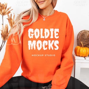 Könnte beinhalten: Orangefarbener Pullover mit dem Text "Goldie Mocks Mockup Studio" in Weiß. Der Pullover wird von einer Person getragen, die auf einem Stuhl sitzt, mit einem Kürbis und einem Weidenkorb im Hintergrund.