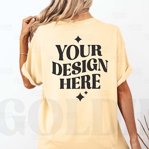 Puede incluir: Una camiseta de color amarillo claro con el texto "YOUR DESIGN HERE" en letras negras. El texto está en un estilo de fuente retro con una forma de diamante arriba y abajo del texto.