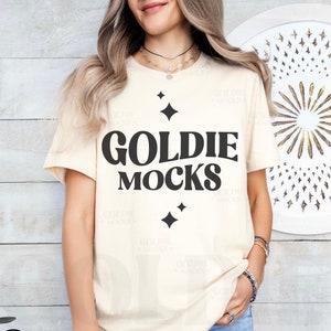 May include: A beige t-shirt with the text "Goldie Mocks" in black lettering. The shirt has a distressed look and features small black diamond shapes.