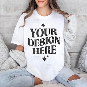 Peut inclure: Un t-shirt blanc avec le texte "YOUR DESIGN HERE" en noir, port&eacute; par une personne assise sur un canap&eacute; avec des coussins gris.