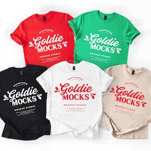 Könnte beinhalten: Fünf T-Shirts in den Farben Rot, Grün, Schwarz, Weiß und Beige. Jedes Shirt zeigt den Text "Goldie Mocks" in einer stilisierten Schriftart, mit zusätzlichem Text darunter. Die Shirts sind vor einem weißen Hintergrund angeordnet.