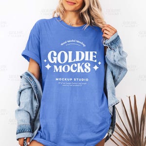 Puede incluir: Una camiseta azul con el texto "GOLDIE MOCKS" en blanco, con una chaqueta vaquera sobre los hombros. La camiseta tiene un corte holgado y la lleva una persona.