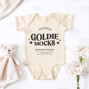 Może przedstawiać: Beżowy śpioszek dla niemowląt z krótkimi rękawami i czarnym nadrukiem z napisem "Real POD Mockup Goldie Mocks Mockup Studio". Śpioszek leży na białym tle z pluszowym misiem i kwiatami.