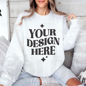 以下が含まれることがあります： 黒い文字で「YOUR DESIGN HERE」と書かれた白いクルーネックのスウェットシャツ。星のアクセント付き。スウェットシャツは、枕のあるソファに座っている人に見せています。