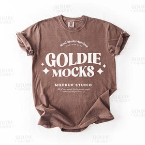 Könnte beinhalten: Ein braunes T-Shirt mit einer weißen Grafik, auf der "GOLDIE MOCKS MOCKUP STUDIO" mit einem weißen Stern auf jeder Seite des Textes steht. Der Text ist in einer Retro-Schriftart.