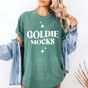以下が含まれることがあります： 白い文字で「Goldie Mocks」と書かれた緑色のTシャツを着た女性。デニムジャケットを着て、長いブロンドの髪をしています。